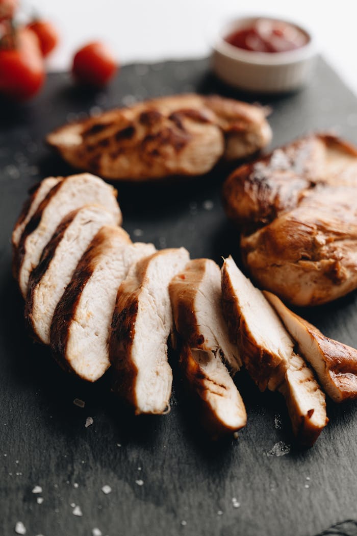 Close-up of grilled chicken breasts sliced on slate with tomato garnish.